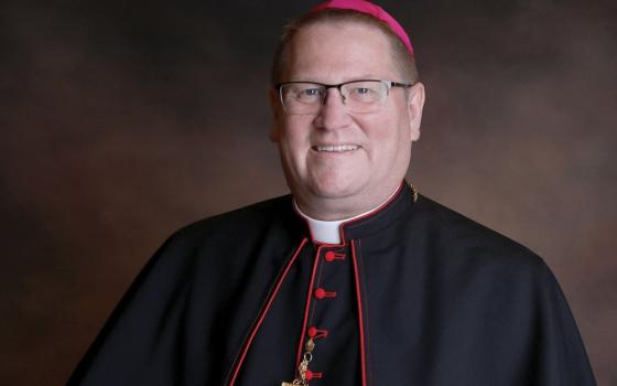 A white man wearing a violet zucchetto, glasses, and a bishop's cassock smiles at the camera