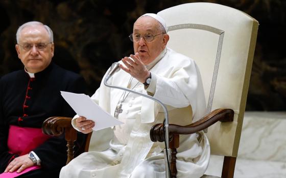 Pope Francis speaks and gestures while sitting in his white chair. A man wearing a bishop's cassock sits to his right.