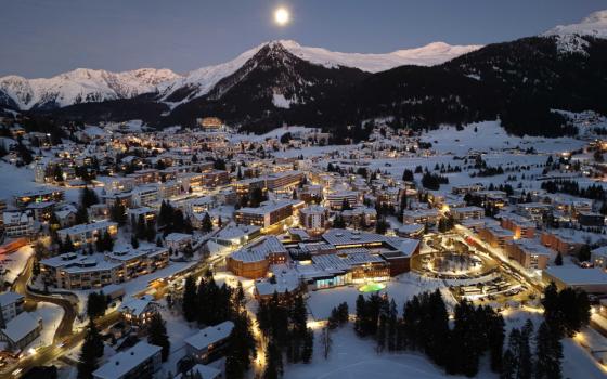 A snowy mountain town seen from above is lit up with warm lights below the full moon