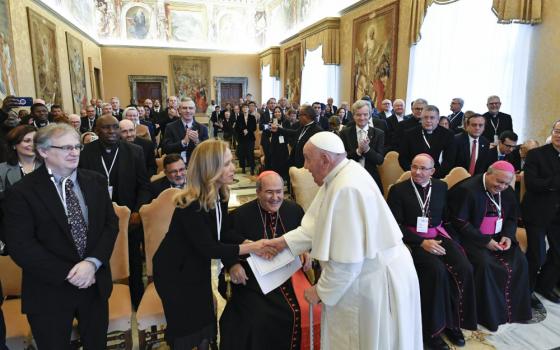Pope Francis shakes hands with a blond woman in business clothes in front of a hall full of people