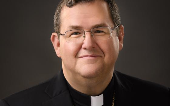A white man with brown hair and glasses smiles at the camera. He wears a clerical collar and a pectoral cross.