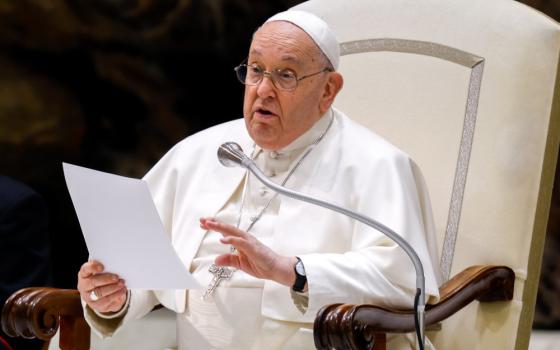 Pope Francis sits in his white chair and speaks into a microphone while holding a piece of paper