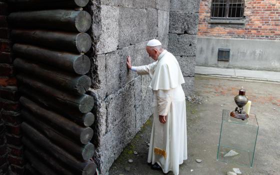 Pope Francis touches a gray wall while bowing his head. He stands in an outdoor space.