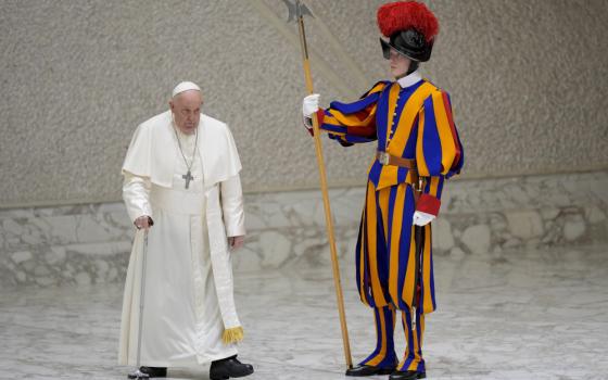 Pope Francis walks with a serious expression past a Swiss Guard wearing his colorful outfit