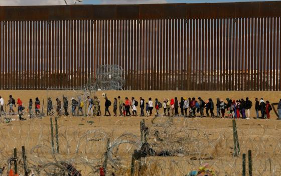 People seeking asylum lined before border wall in background.