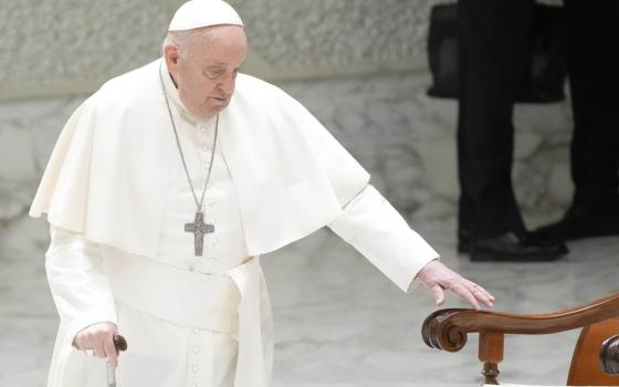 Pope Francis walks with cane towards chair. 