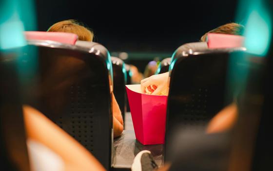 View of movie theater seats from behind, as one patron reaches for popcorn (Unsplash/Jakub Zerdzicki)
