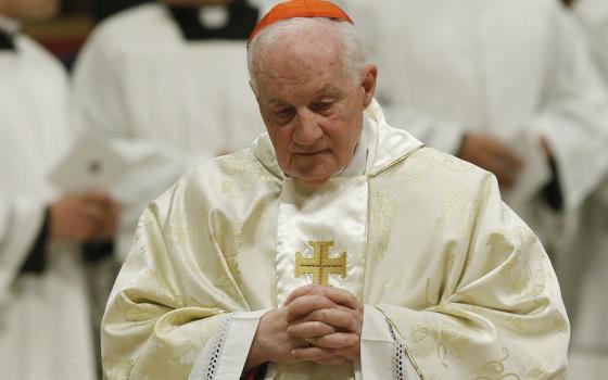 Prelate, vested, bows head and folds hands in prayer. 