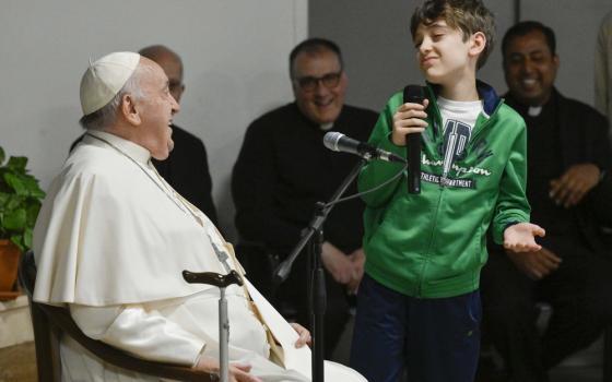 Child stands, with microphone, to ask Pope Francis, seated, a question; all smile. 
