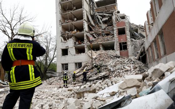 A first-responder, standing on rubble, faces a bombed-out building.