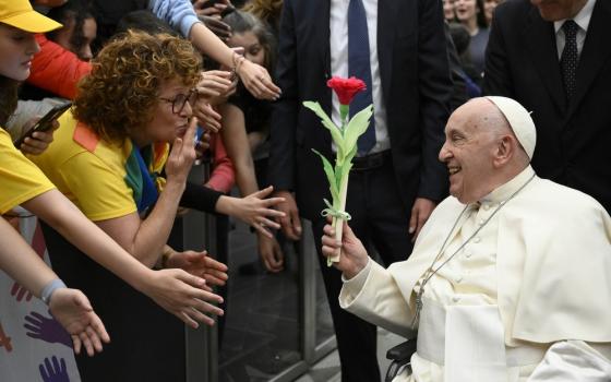 Pope Francis, in wheelchair, holds flower, and smiles at woman who blows kiss from behind barrier.