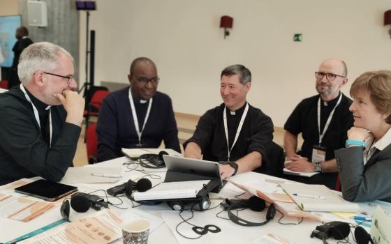 Group of priest sit around table, along with sister, smiling and talking. 