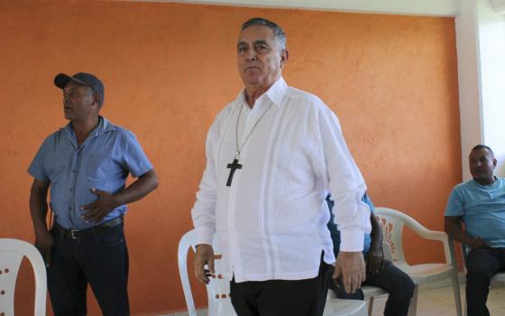 Bishop wears plain white button-down shirt and wooden cross, while entering room. 