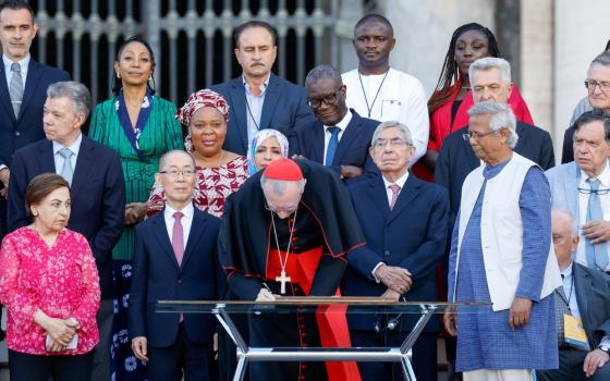 Cardinal bends over table to sign document, with others in background. 