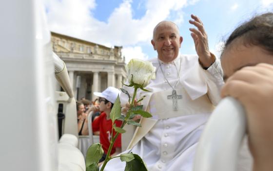 Pope Francis, seated, framed by faces and a white rose, as he smiles and raises his hand in blessing. 