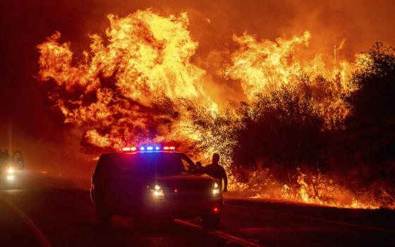 Towering conflagration pierces night sky, emergency vehicle seen in foreground