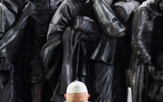 Pope Francis, seated, bows head in prayer, with bronze procession of migrants directly behind. 