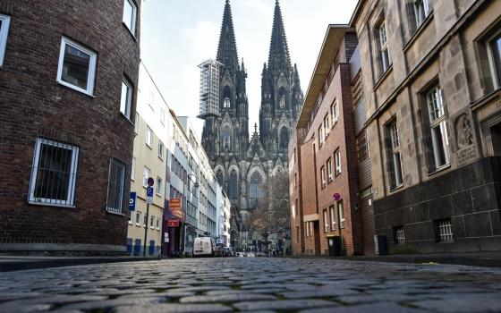 Cathedral of Cologne, one spire obscured by scaffolding, looms over cobblestone street.
