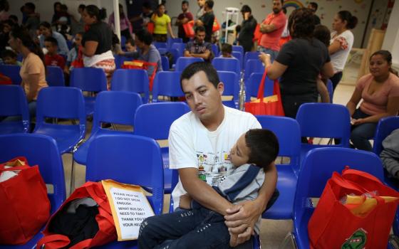 Man, holding child, sits in blue plastic chair. Room behind him full. 