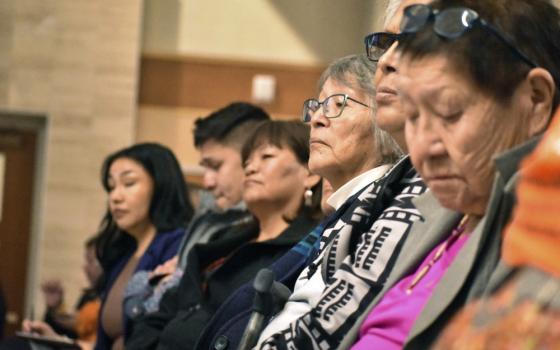 A long line of seated people are shown from the side, their faces somber. 