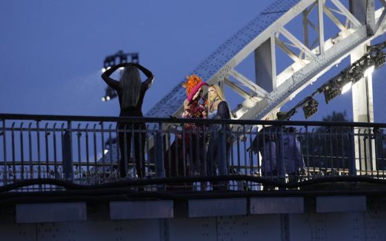 Drag queen on bridge at night, backlit by bridge's arch. 