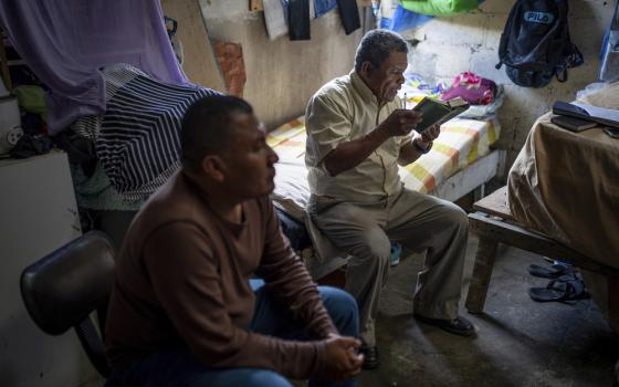 Alvicio sits on a twin bed in a cinderblock room reading from bible, Ocampo sits to his right on a chair.