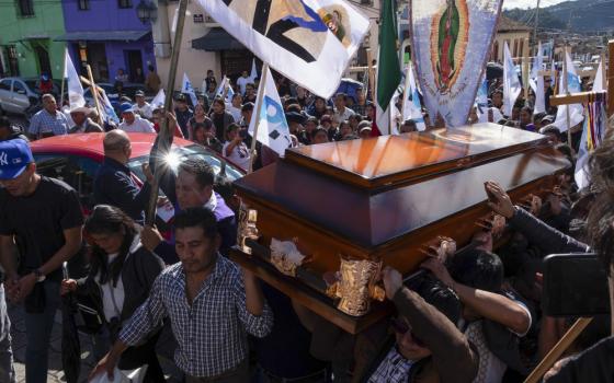 Pall-bearers carry casket, surrounded by large group carrying banners and votives.