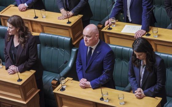Prime Minister, wearing blue suit, stands with folded arms, as others around him bow their head solemnly.