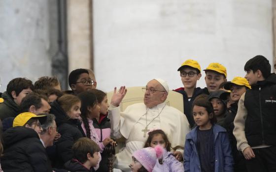 Pope sits surrounded by children; he gestures and smiles, wearing glasses.