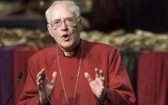 Carey shown with hands mid-gesture while speaking, wearing red cassock.