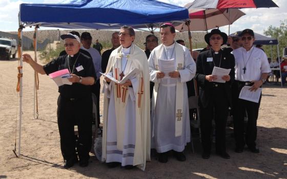 The archbishop wears white vestments and reads from book, others assembled around under tarpaulin tent and umbrellas.