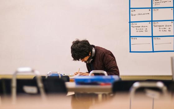 A young man works in a classroom setting. He is seated amid tables and chairs, with a dry erase board behind him. (Unsplash/Jeswin Thomas)