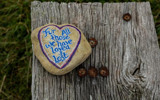 A rock with "For all those we have lost and lost" painted on it. The rock is sitting on a wooden bench. (Unsplash/Nick Fewings)