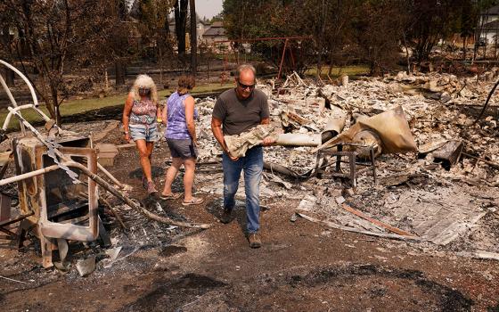 Tim Hansen carries a statue of an angel retrieved from the debris as his wife, Shelly, and Nancy Fuller survey their destroyed home in Spokane, Wash., Aug. 3, 2026, after a wildfire swept through the neighborhood. (OSV News/Reuters/David Ryder)