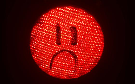 A photo illustration shows a circular red light with a drawing of a sad face on it, against a dark black background. (Unsplash/Filipe Nobre)