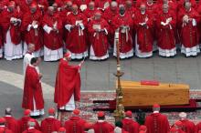 Many cardinals wearing red surround a wooden casket on a rug in St. Peter's Square