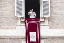 Pope Francis raises his hand while standing in a window behind a clear podium