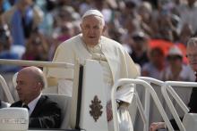 Pope Francis stands in the back of the popemobile. A crowd is visible and blurred behind him.