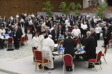 Pope Francis stands at a round table with clerics and laypeople. Other round tables are visible throughout the hall.