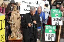 Bishop Strickland holds a rosary. People around him are bowing their heads. A person behind him holds a sign that reads: "We stand with Bishop Strickland."